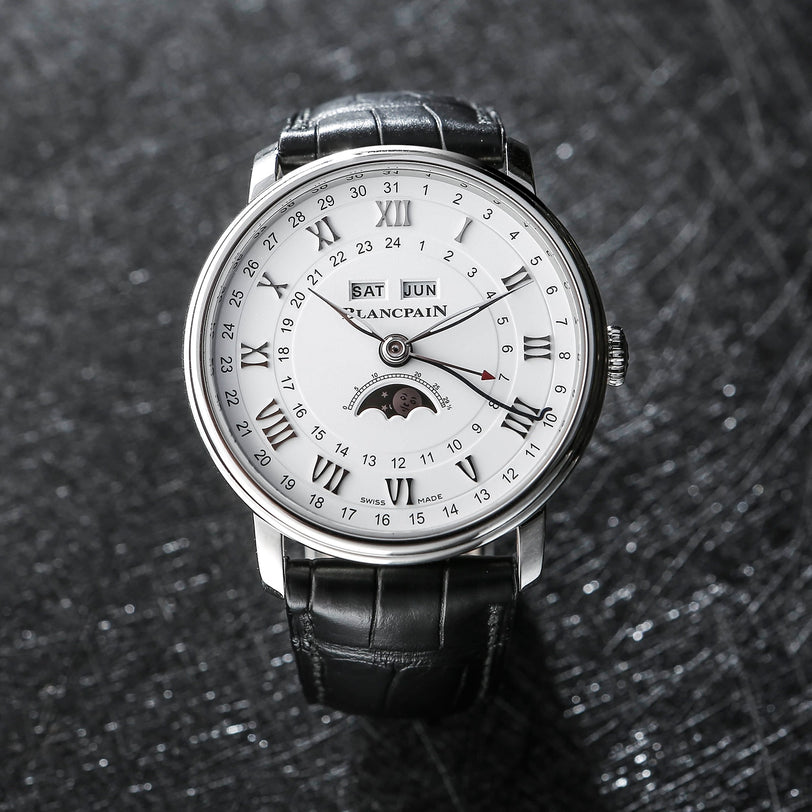 A Blancpain Villeret Quantième Complet wristwatch features a black leather strap, Roman numerals, moon phase, and complete calendar with day, date, and month indicators on a dark textured dial.