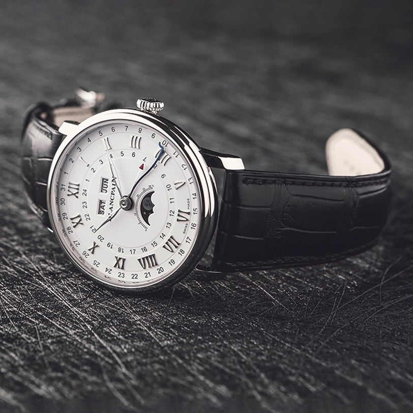 The Blancpain Villeret Quantième Complet, featuring a white dial with Roman numerals, moon phase display, complete calendar, black leather strap, and multiple subdials, rests elegantly on a dark textured surface.