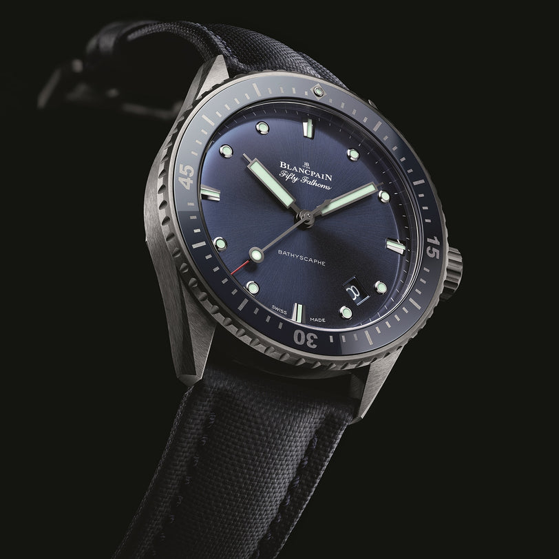 The Blancpain Fifty Fathoms Bathyscaphe features a blue dial, luminous markers, automatic movement, and a black fabric strap, shown on a black background.