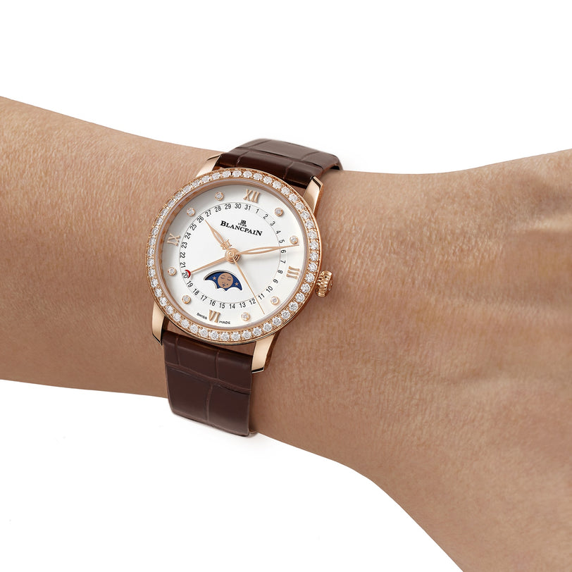 A wrist wearing the Blancpain Villeret Quantième Phases de Lune, featuring a brown leather strap, diamond bezel, Roman numerals, and an elegant moon phase on its 18K red gold dial.