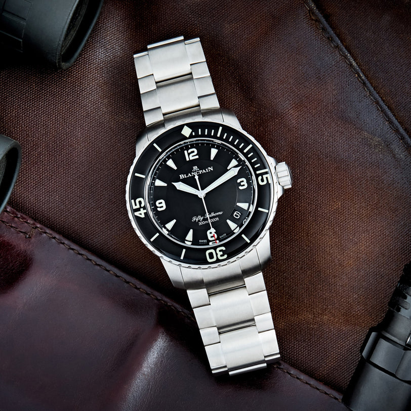 The Blancpain Fifty Fathoms Automatique is a stainless steel diver’s watch with a black dial and bezel, shown on brown leather and also available with a metal bracelet.