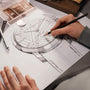 An artist sketches a detailed Vacheron Constantin Patrimony Moon Phase Retrograde Date design on paper, surrounded by drawing tools and a precision moon phase watch face component.