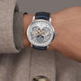 Wearing a beige coat, a person showcases the Vacheron Constantin Traditionnelle Complete Calendar Openface— a limited edition platinum watch with blue leather strap, exposed gears, and calendar displaying day, date, and moon phase.