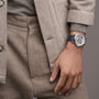 A person in a beige suit with a button-up coat and trousers, wearing a Vacheron Constantin Traditionnelle Complete Calendar Openface platinum watch with a black leather strap.