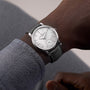 A close-up of a person wearing a Vacheron Constantin Traditionnelle Manual-Winding wristwatch featuring a white dial and gray leather strap.