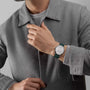 A person in a gray coat and sweater adjusts their collar, displaying a Vacheron Constantin Patrimony Self-Winding 18K white gold watch with a dark strap and Geneva Seal on their left wrist.
