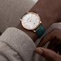 A close-up of a wrist with dark skin in a light gray sleeve wearing the Vacheron Constantin Patrimony Self-Winding, a limited edition gold watch featuring a white face, date display, Geneva Seal, and green leather strap.