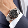 A person wearing a Breitling Superocean Heritage B20 Automatic 42 with a black dial and mesh strap on their left wrist, dressed in a dark blue suit jacket and white shirt.