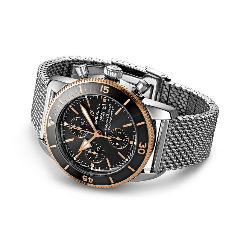 The Breitling Superocean Heritage B20 Automatic 44 is a 44mm stainless steel chronograph watch with a black dial, gold bezel, mesh bracelet, water resistance, and multiple subdials displayed on a white background.