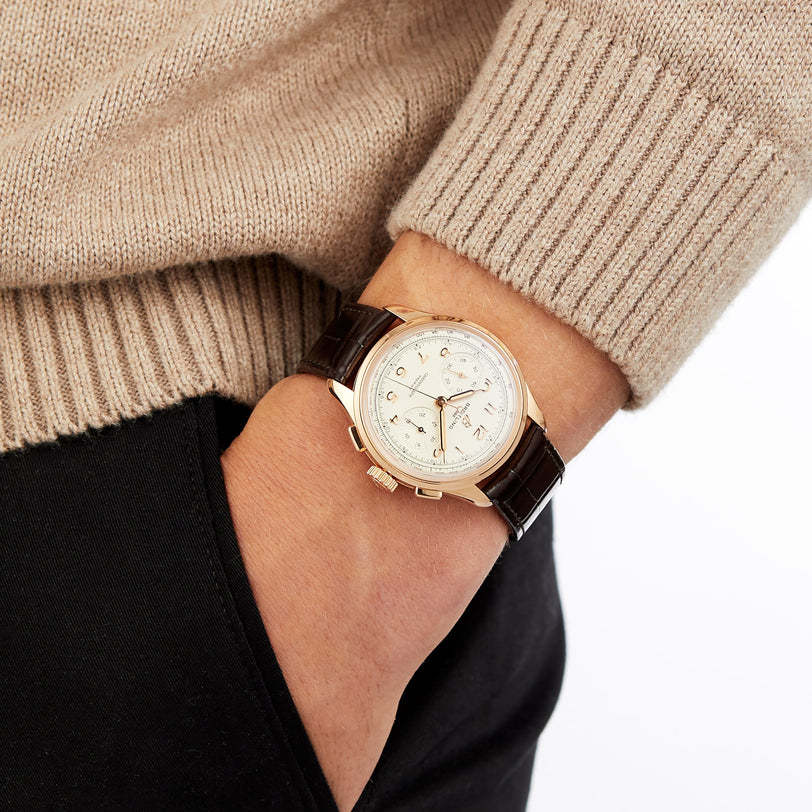 A person in a beige sweater and black pants has their left hand in their pocket, showcasing a Breitling Premier B09 Chronograph 40 wristwatch with a brown leather strap and white dial.