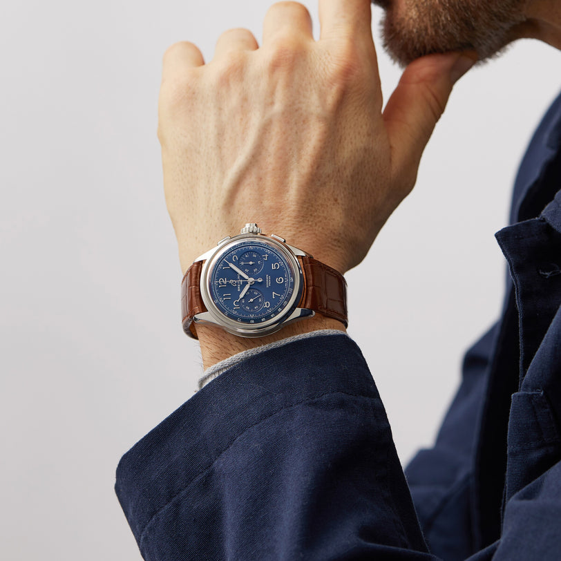 A person wearing a blue jacket and a Breitling Premier B15 Duograph 42 with a brown leather strap and blue dial rests their chin on their hand.