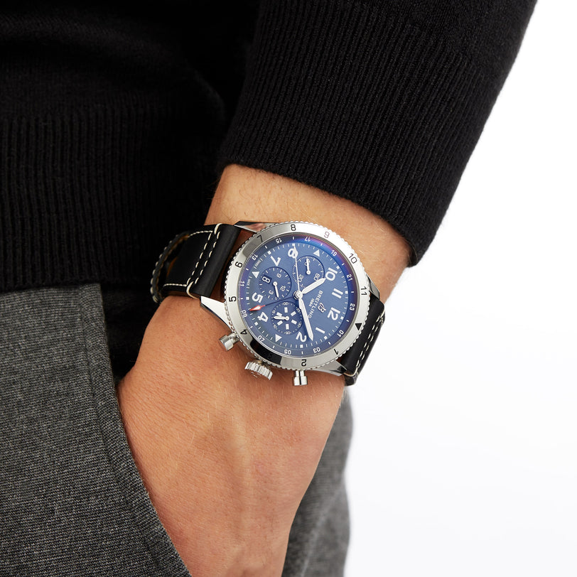 A person in a black long-sleeve shirt and grey pants wears a Breitling Super AVI B04 Chronograph GMT 46 Tribute To Vought F4U with a blue dial and black leather strap on their left wrist.