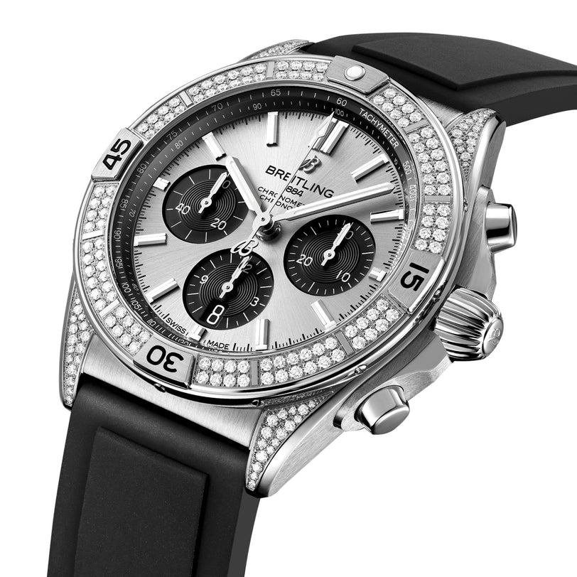 The Breitling Chronomat B01 42 is a silver chronograph watch featuring a diamond bezel, three subdials, date display, stainless steel case, and a black rubber strap.