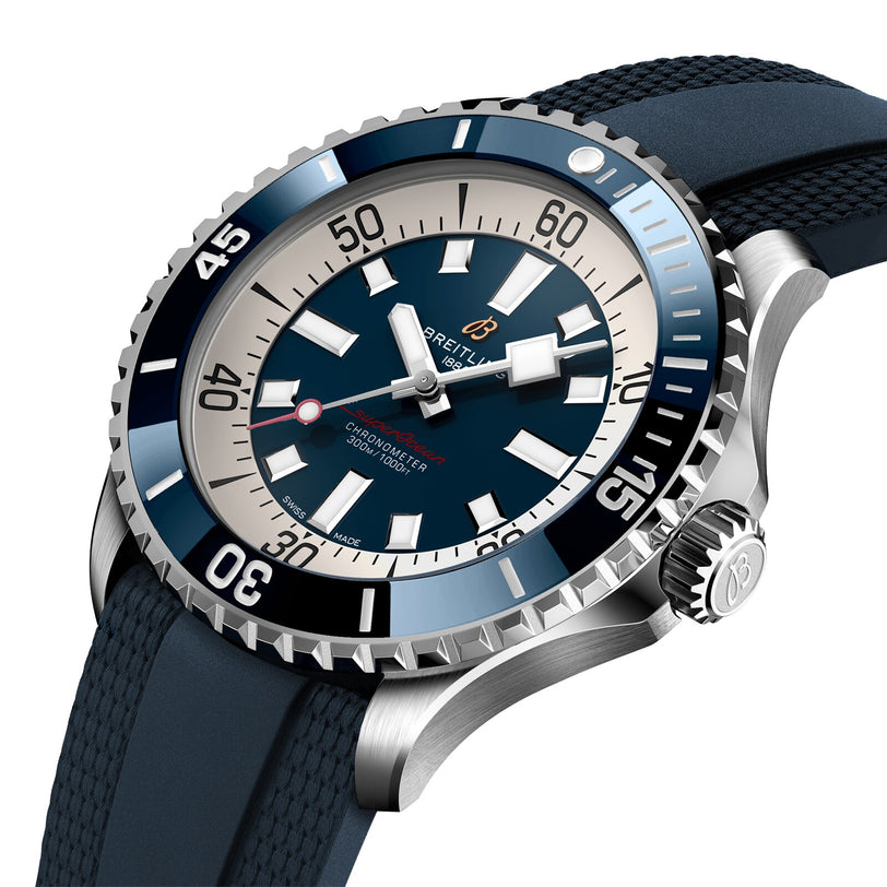 The Breitling Superocean Automatic 46 is a stainless steel dive watch with a blue bezel, cream and blue dial, date window, blue rubber strap, and water resistance. Photographed at an angle.