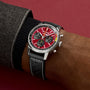 A person wearing the Breitling Top Time B01 Chevrolet Corvette watch—with a red dial, three subdials, black leather strap, and silver case—stands against a burgundy background.