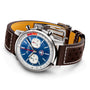 The Breitling Top Time B01 Shelby Cobra chronograph features a blue dial, two subdials, brown perforated leather strap, silver case, and Manufacture Calibre 01, displayed at an angle on a white background.