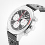A close-up of the Breitling Top Time B01 Deus chronograph, featuring a black and white dial with three subdials, date window, black leather strap with white stitching, and the Breitling Manufacture Calibre 01 movement.