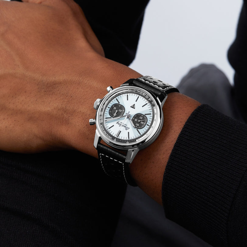 A person in a black long-sleeve shirt wears a silver and black Breitling Top Time B01 Triumph chronograph with a white dial and black subdials.
