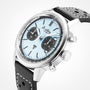A close-up of the Breitling Top Time B01 Triumph shows its stainless steel case, light blue dial with black subdials, and a black leather strap with white stitching—powered by the Breitling 01 calibre against a plain background.