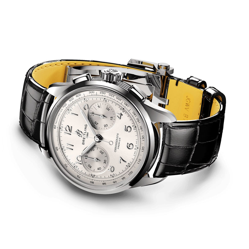 The Breitling Premier B09 Chronograph 42 features a silver stainless steel case, white dial with three subdials, black Arabic numerals, and a black leather strap with yellow underside.