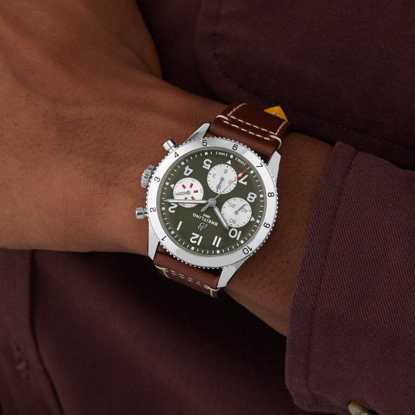 A person in a brown shirt wears a Breitling Classic AVI Chronograph 42 Curtiss Warhawk with a brown leather strap, green dial, and three subdials on their left wrist.