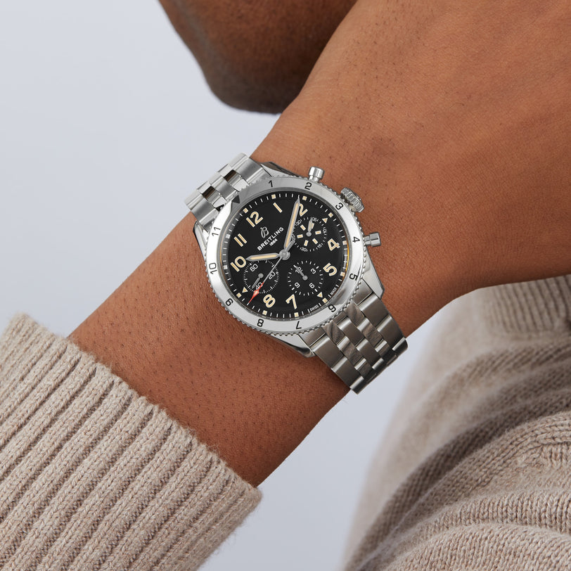 A person wearing the Breitling Classic AVI Chronograph 42 P-51 Mustang, a silver pilot’s watch with a black dial and yellow numerals, inspired by aviation heritage, paired with a beige knit sweater.