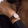 A person with curly hair wears a Breitling Navitimer 32 wristwatch featuring a brown leather strap, white dial, and diamond hour markers on their left wrist.