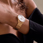 A person wearing an off-the-shoulder black top and a Breitling Navitimer 32 gold wristwatch featuring a mother-of-pearl dial and diamond hour markers.