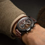 A close-up of a person wearing the Breitling Top Time B21 Chevrolet Corvette—this brown racing chronograph features an open-heart mechanism, brown strap, and peeks from a beige jacket sleeve; ideal for classic sports car enthusiasts.