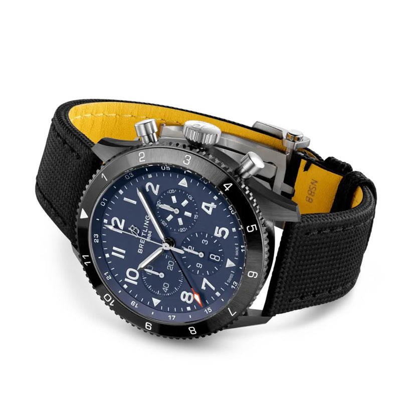 A black Breitling Super AVI B04 Chronograph GMT 46 Tribute To Vought F4U with a blue dial, numbered bezel, and black fabric strap with yellow lining, shown on a white background.