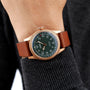A person wears an Oris Big Crown Pointer Date 80th Anniversary Edition with a green dial, gold case, brown leather strap, and Arabic numerals over a dark long-sleeve top.