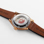 A wristwatch with a brown leather strap is shown face down, revealing the transparent case back and intricate mechanical movement of the Oris Big Crown Pointer Date 80th Anniversary Edition.