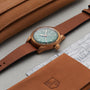 The Oris Big Crown Pointer Date 80th Anniversary Edition wristwatch with a green dial and brown leather strap is shown on a light surface next to tan leather cases, a map fragment, and visible Oris branding.