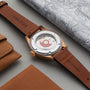 The Oris Big Crown Pointer Date 80th Anniversary Edition, featuring a green dial and brown leather strap, rests on a grey surface beside a leather wallet and map.