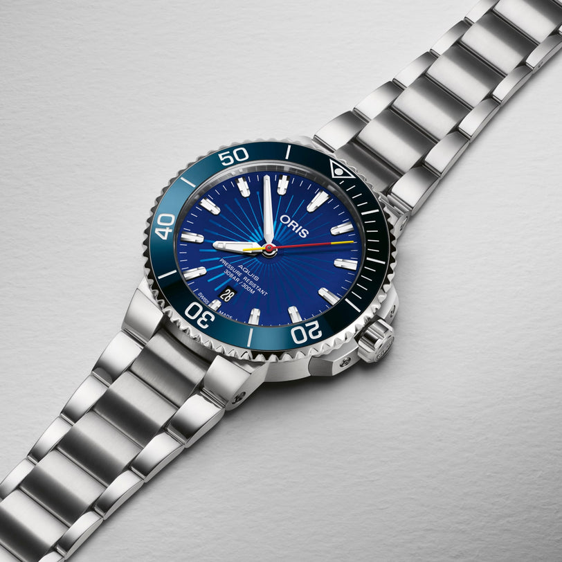 An Oris Aquis Sun Wukong Limited Edition wristwatch with a blue dial, luminescent markers, rotating bezel, and metal bracelet is displayed on a light gray textured surface.