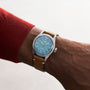 A person wearing an Oris Big Crown Oris X Cervo Volante watch with a light blue dial and brown leather strap, paired with a red long-sleeve top.
