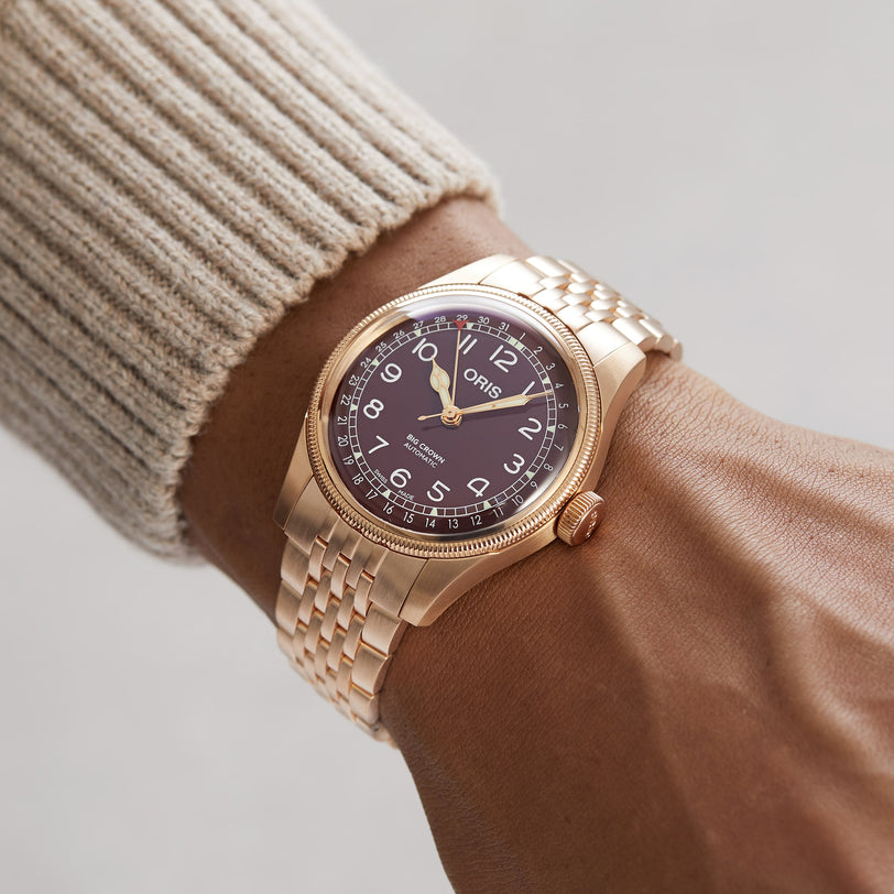 An Oris Big Crown Bronze Pointer Date wristwatch with a brown dial and metal band worn on a person’s wrist in a beige sweater.