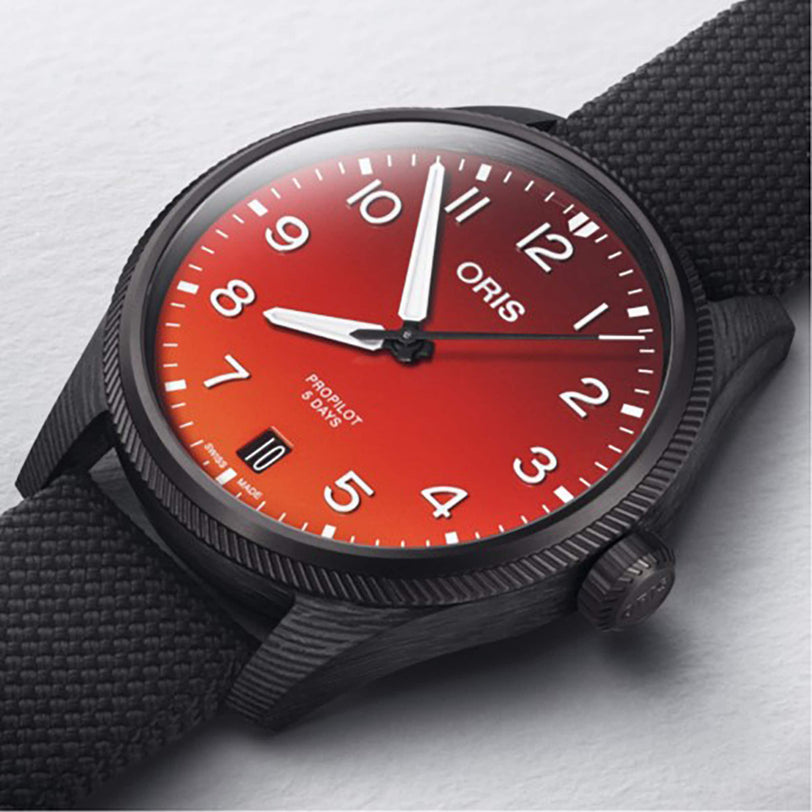 The Oris ProPilot Coulson Limited Edition features a textured black strap, gradient red dial with white numerals and hands, and a 3D printed carbon fibre case, all inspired by Coulson Aviation and displayed on a light surface.
