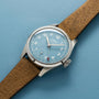 The Oris Big Crown Pointer Date Calibre 473, with a light blue dial, white numerals, small seconds subdial, and brown leather strap, showcases the hand-wound calibre 473 and rests on a matching blue surface for timeless style.
