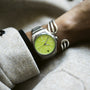 A person wearing an Oris ProPilot X Kermit Edition with a vibrant green dial and silver bracelet on their wrist, paired with a light coat.