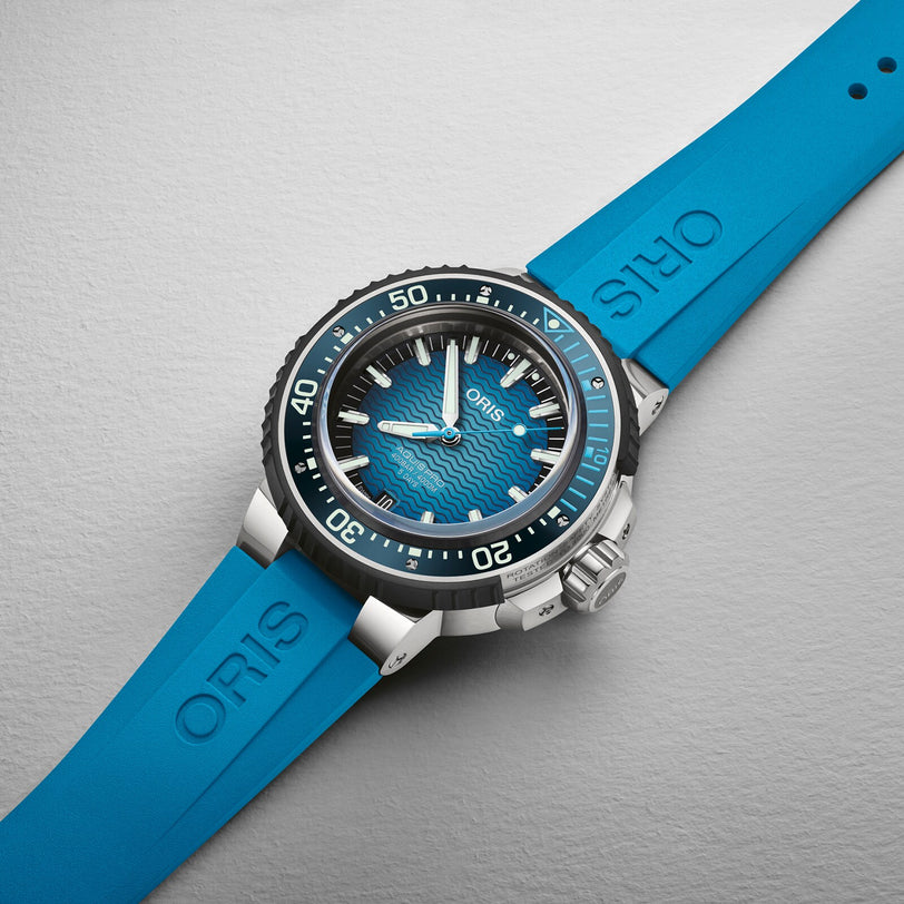The Oris Aquis Pro 4000M dive watch features a blue wave-patterned dial, black bezel, blue rubber strap with Rotation Safety System, and is displayed on a light gray surface.