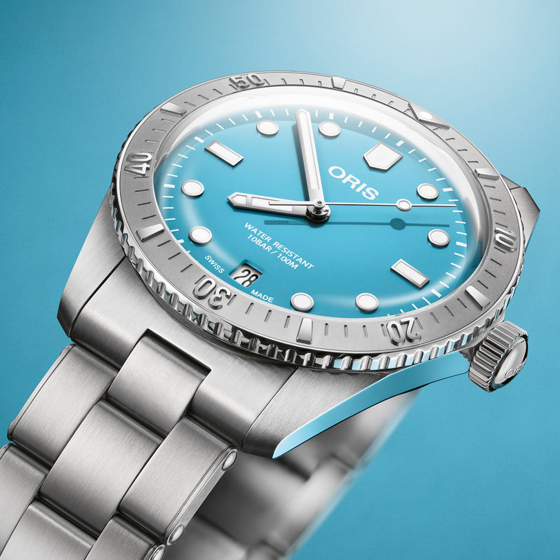 The Oris Divers Sixty-Five Cotton Candy wristwatch features a silver case, turquoise dial, and metal bracelet, displayed on a blue background.