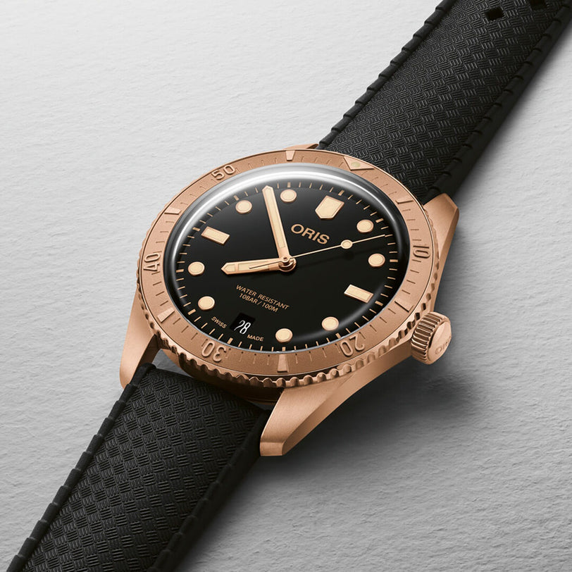 The Oris Divers Sixty‑Five Date wristwatch features a gold case, black dial, and textured black strap, displayed on a light gray background.