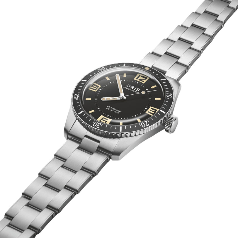 The Oris 60th Anniversary Edition is a stainless steel dive watch with a black dial, bold numeral markers, and metal link bracelet, powered by a Swiss Made automatic movement and shown at an angle on a white background.