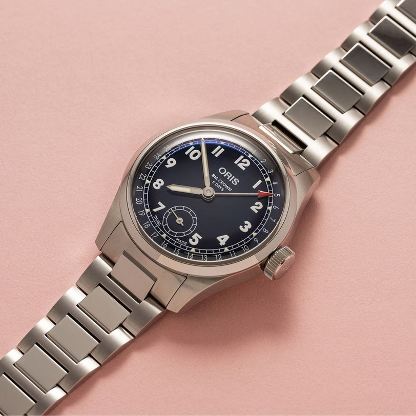 The Oris Big Crown Pointer Date Calibre 403, featuring a stainless steel case, midnight blue dial with Arabic numerals, and metal link bracelet, is elegantly displayed on a light pink background.