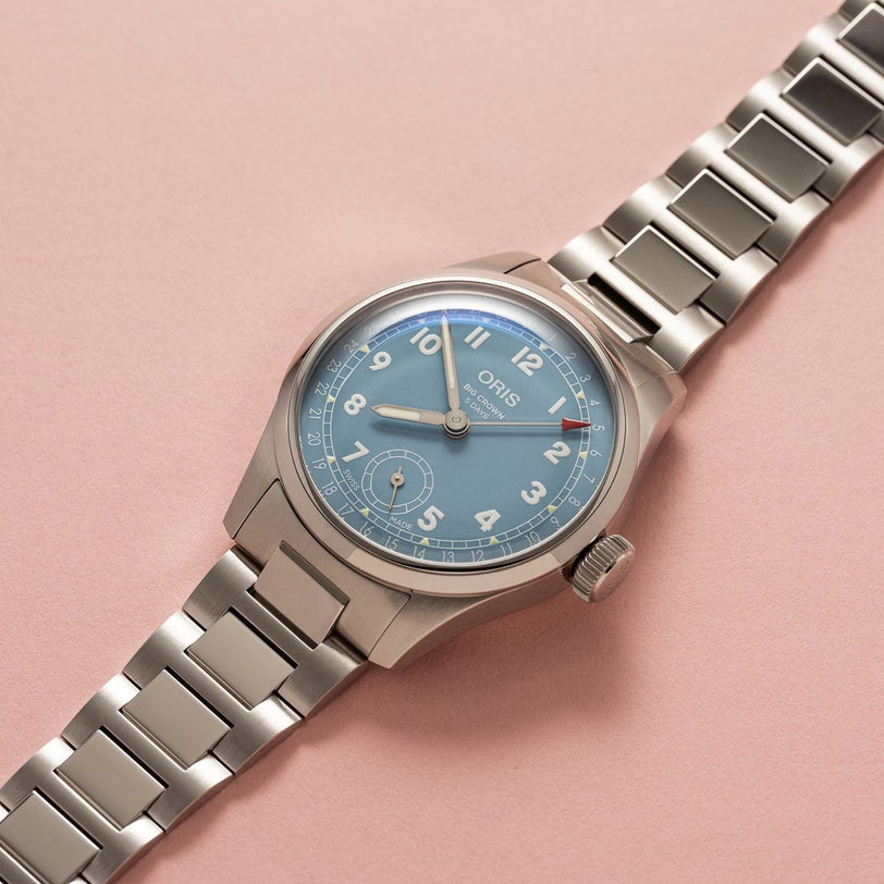 The Oris Big Crown Pointer Date Calibre 473, a stainless steel watch with a light teal blue dial, silver hour markers, and a metal bracelet, is shown on a light pink surface.