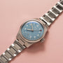 The Oris Big Crown Pointer Date Calibre 473, a stainless steel watch with a light teal blue dial, silver hour markers, and a metal bracelet, is shown on a light pink surface.