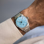 A wrist wears the Oris Big Crown Pointer Date Calibre 473, featuring a silver case, light teal blue dial, silver hour markers, and a metal band, set against a neutral background.