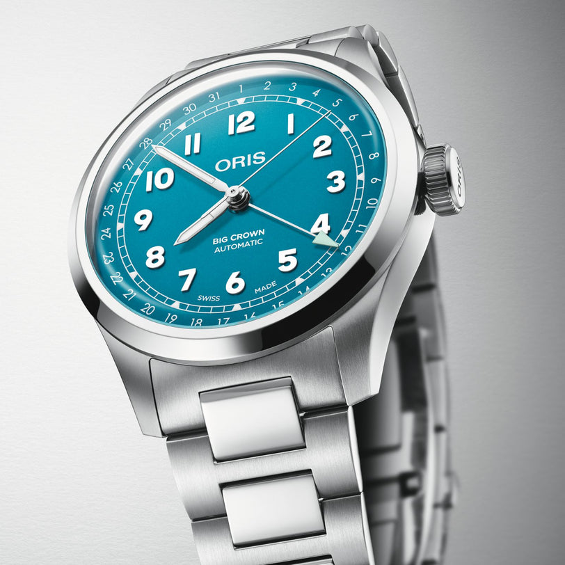 The Oris Big Crown Pointer Date wristwatch, featuring a turquoise blue dial with white numerals and a stainless steel link bracelet, is displayed against a neutral background.