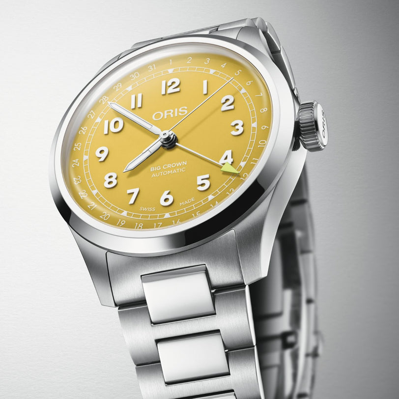 A close-up highlights the Oris Big Crown Pointer Date watch with a sunshine yellow dial, white numerals, and stainless steel bracelet, showcasing its Swiss Made automatic mechanical movement against a neutral background.